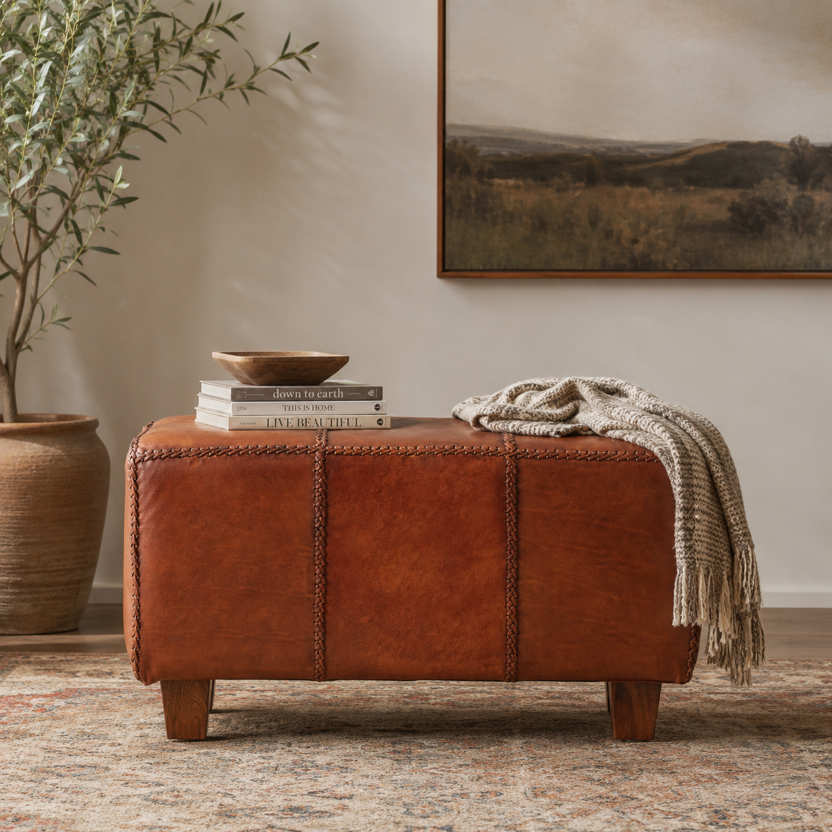 Tierra Leather Bench With Teak Wood Legs styled with books and a rug on top, an olive tree in the corner and artwork on the wall behind
