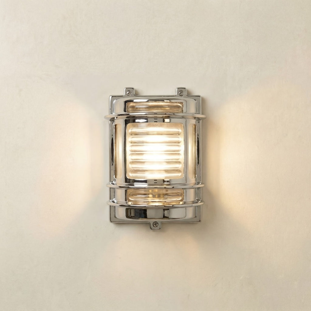 A rectangular wall-mounted light fixture with a polished chrome frame and a warm glowing light inside, mounted on a textured off-white wall.