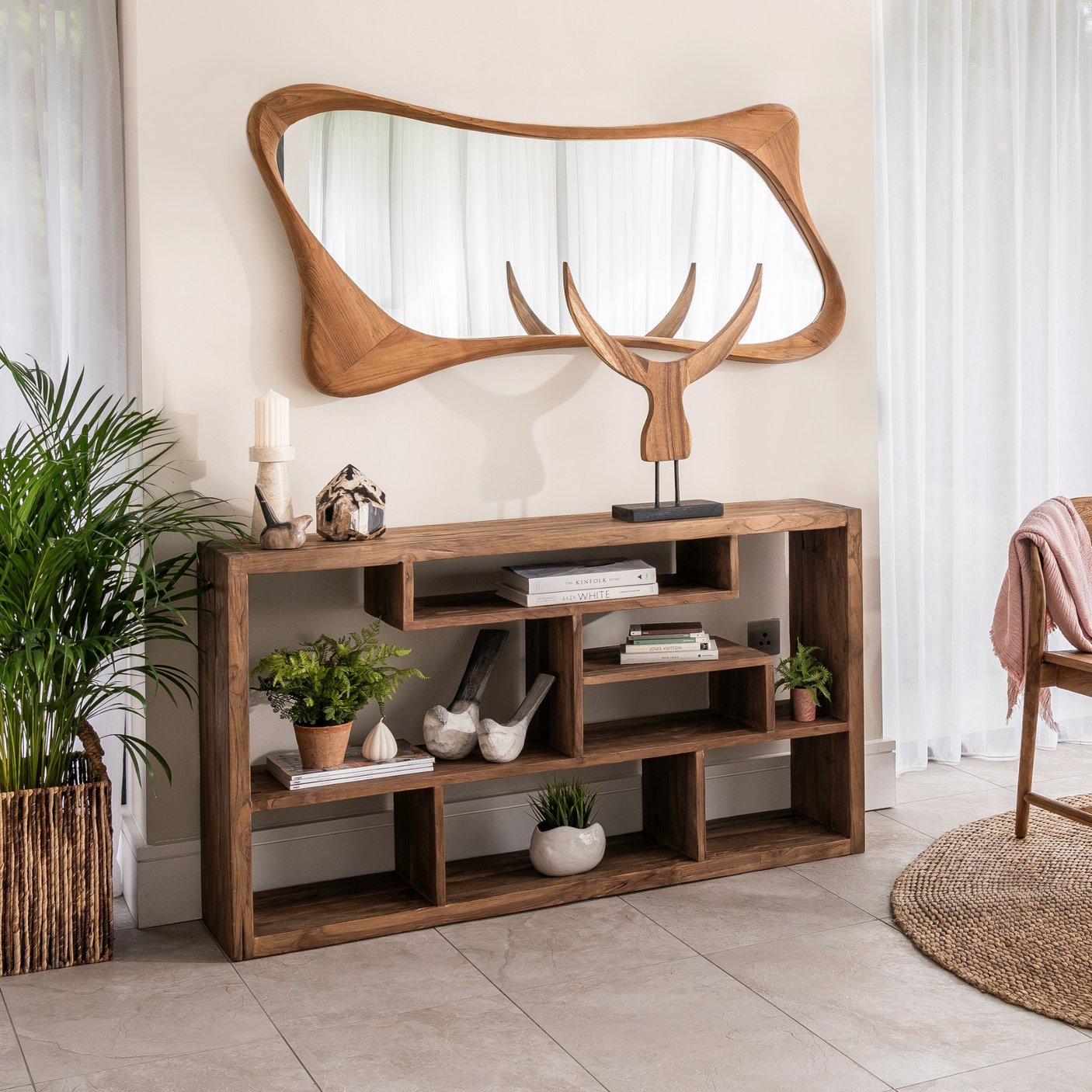A rustic brown wooden console table with open shelving, styled in a bright room with a matching organic-shaped wooden wall mirror above it.