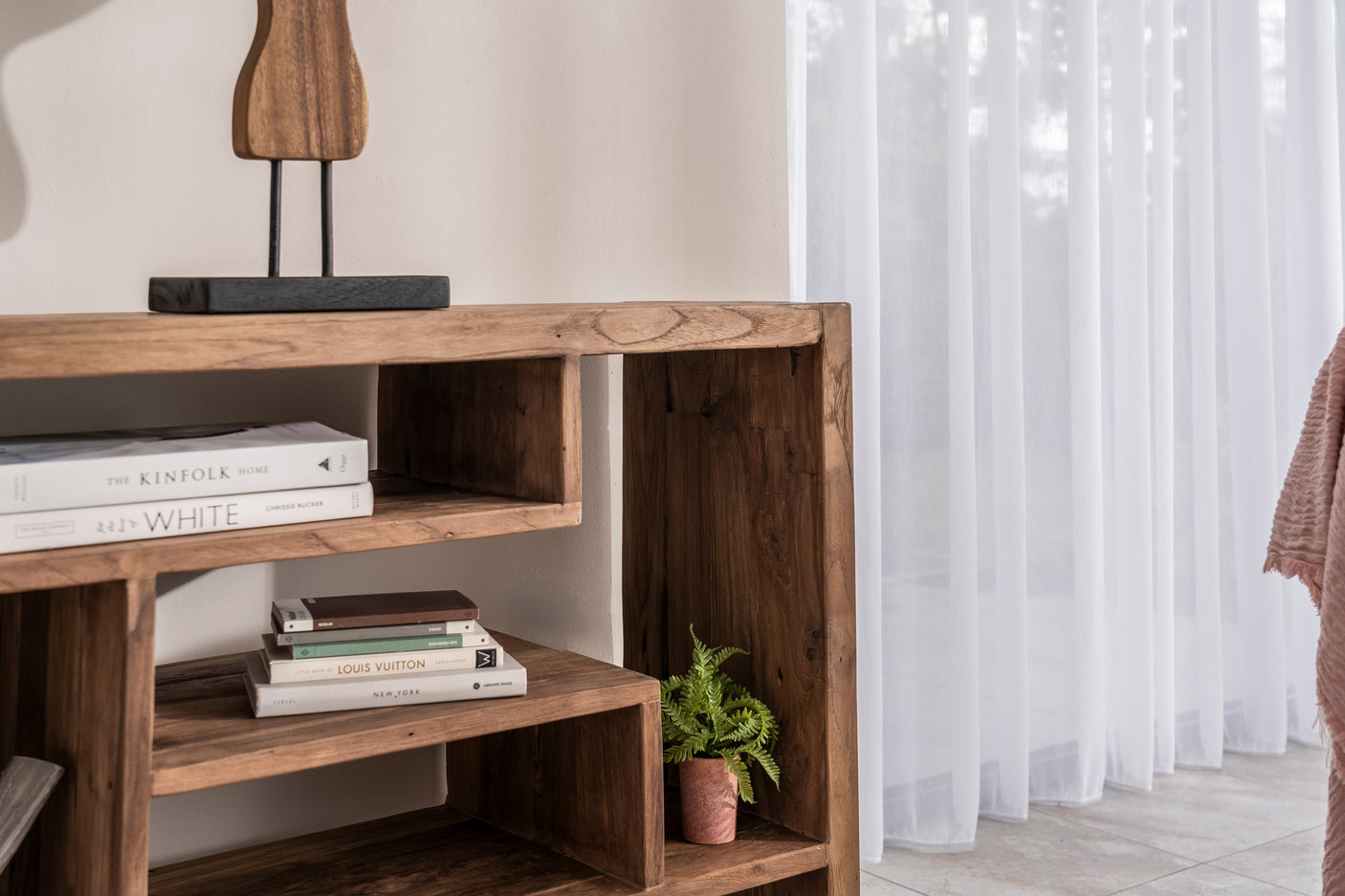 A rustic dark wood open shelving unit styled with books, a small potted fern, and a decorative lamp, set against a neutral wall with white sheer curtains.