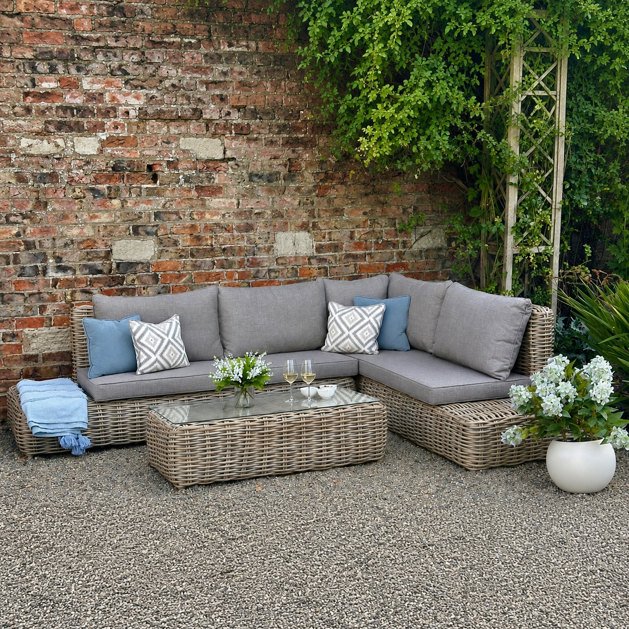 Outdoor furniture set arranged in a corner, consisting of a sofa with cushions and a coffee table, made of wicker, on a patio with a brick wall in the background.