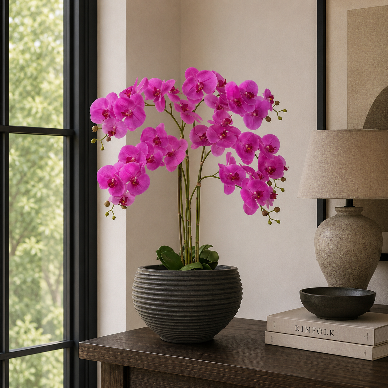 Pink orchid in a dark pot on a wooden table with books and a lamp in a room.