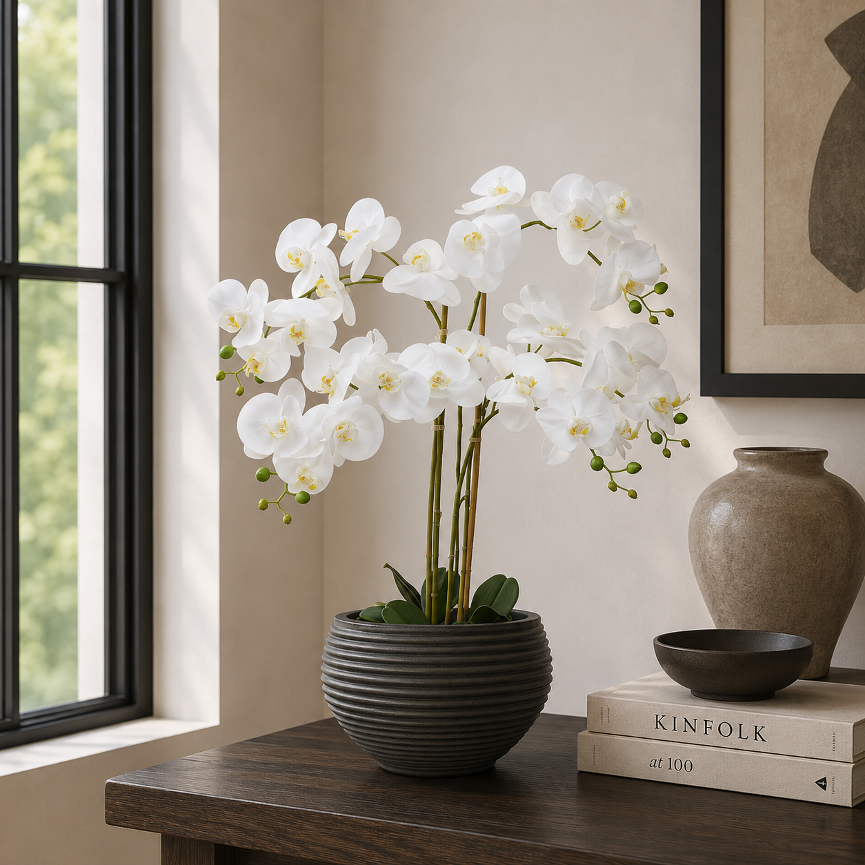 White orchid in a black pot on a wooden table with books and a vase in the background.