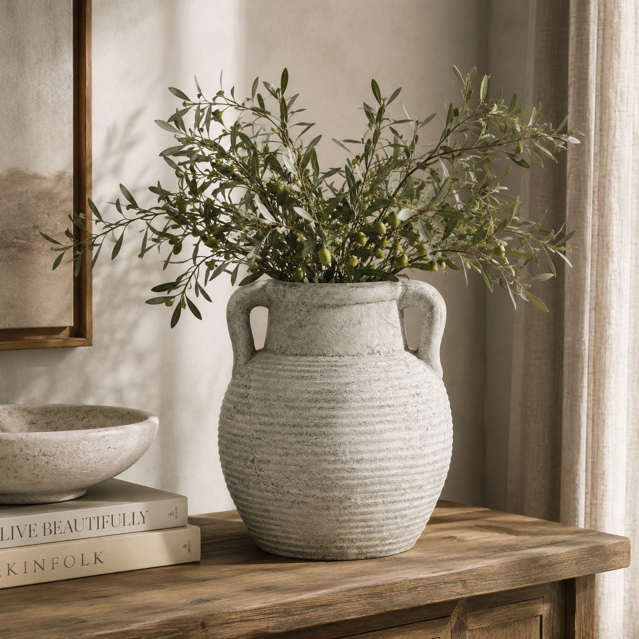 Decorative vase with greenery on a wooden surface with books and a framed picture in the background.