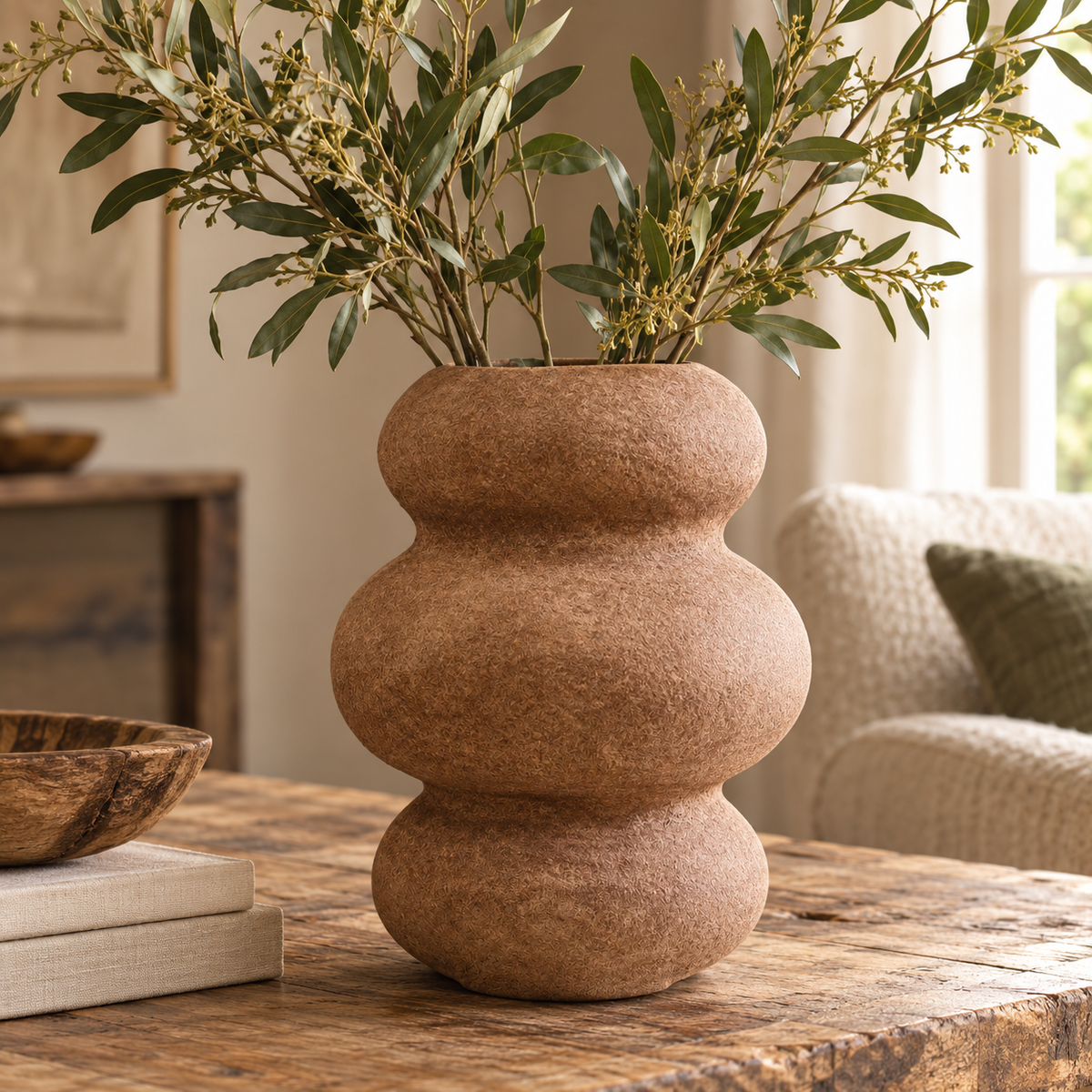 Avara Cotton Mache And Coco Shell Bubble Vase holding leafy green stems on a rustic wooden table, beside books and a wooden bowl, with an armchair behind
