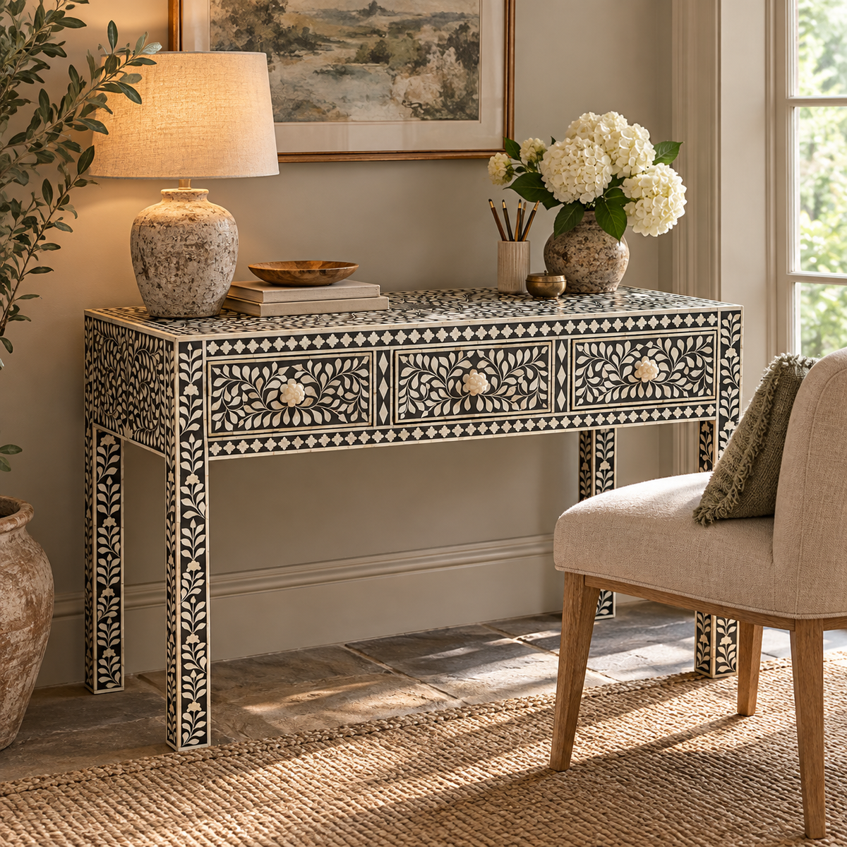 A bone-inlay console table styled in a warm, neutral-toned room with a table lamp, flowers, and a chair.