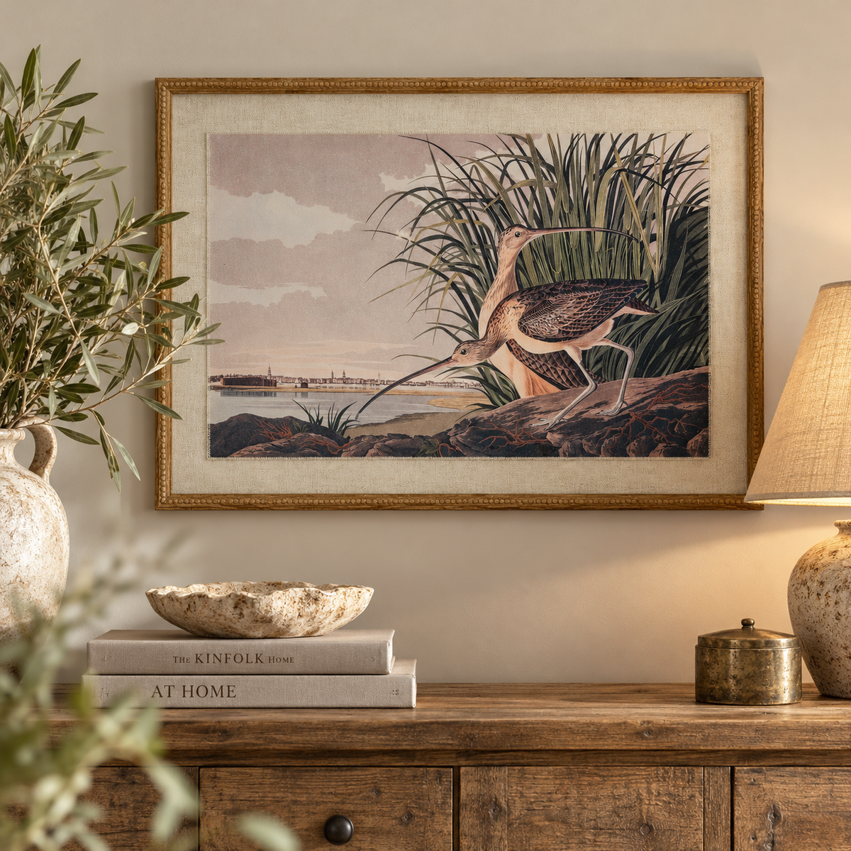 Flora & Fauna Curlews print in a beaded oak-finish frame above a rustic wooden sideboard with olive branches in a stone jug, a linen table lamp and books