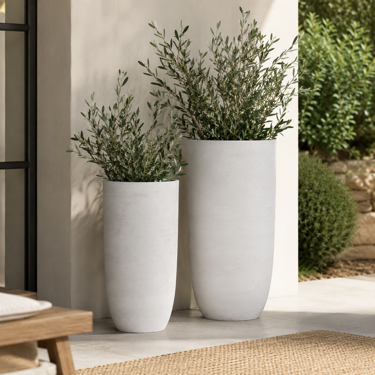 Two tall, cylindrical concrete planters in light gray, filled with green leafy plants, set against a neutral outdoor wall.