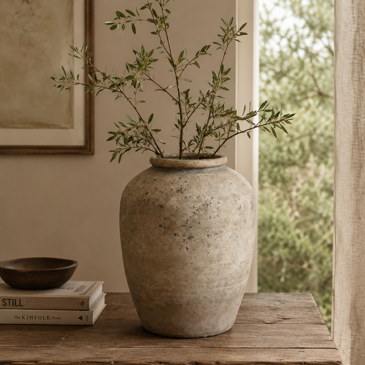 Large terracotta vase with green branches on a wooden surface, with a window and painting in the background.