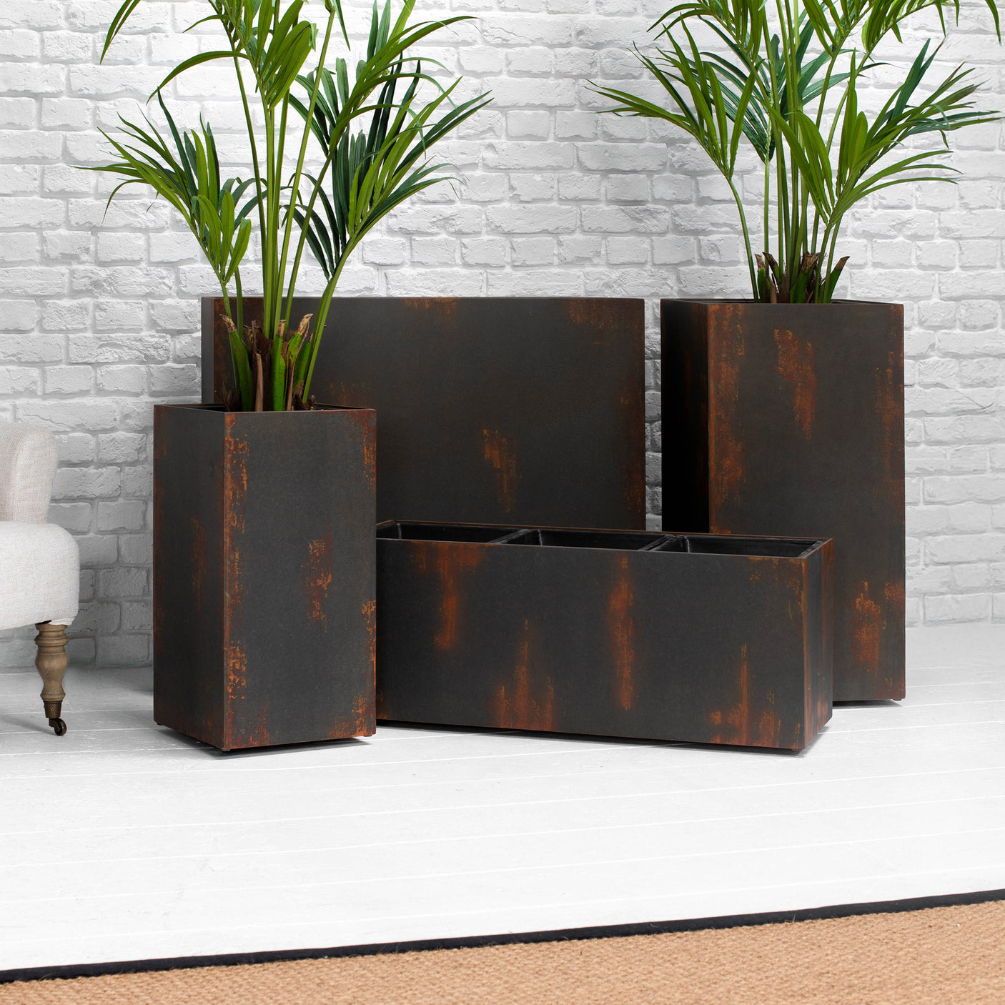 A set of four rectangular metal planters with a distressed rust finish, arranged on a white floor against a white brick wall, with two green palm plants placed inside them.