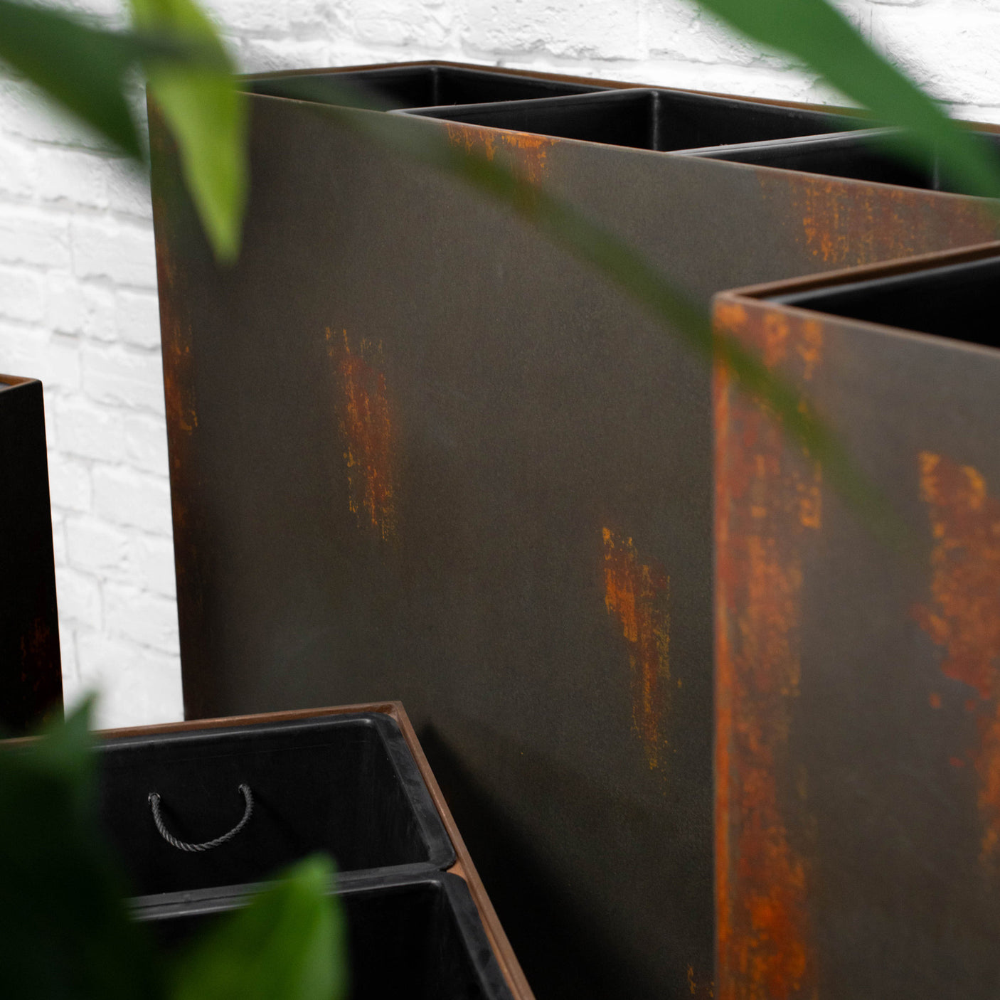 Rectangular metal planter boxes with a dark charcoal finish and rusted orange-brown weathered accents, shown in an outdoor setting with green leaves in the foreground.