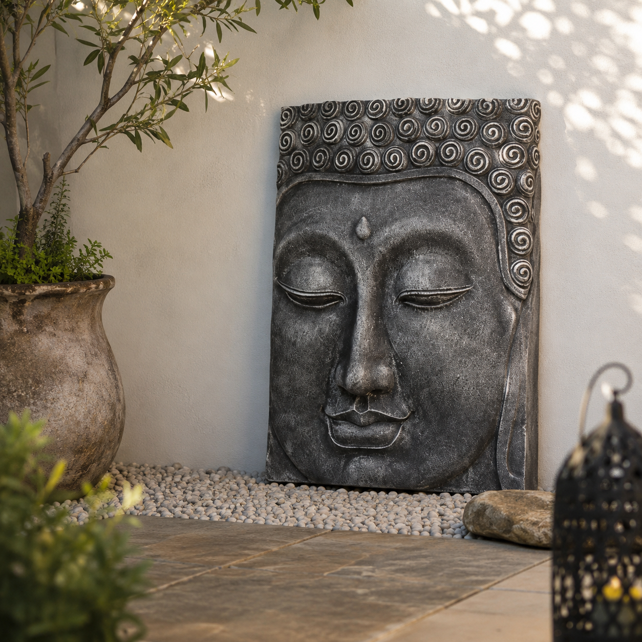 A rectangular stone Buddha head wall sculpture with a dark grey textured finish, displayed in a garden setting against a light beige wall with pebbles, a potted plant, and a decorative lantern.