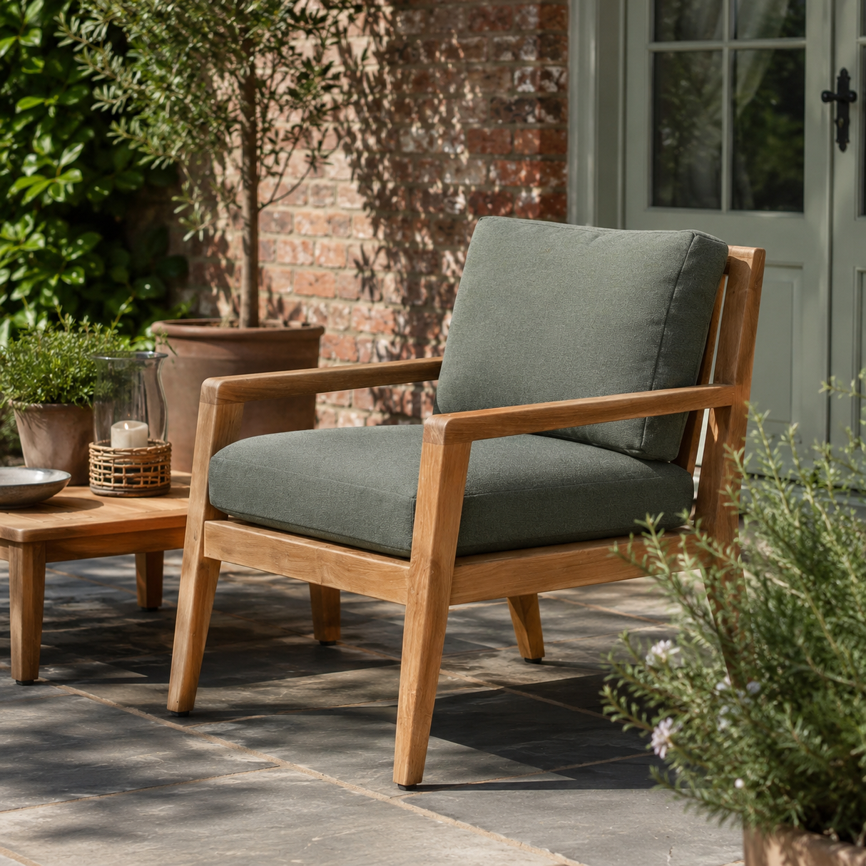 A wooden outdoor armchair with thick sage green cushions sits on a stone patio in a garden setting next to a matching wooden side table.