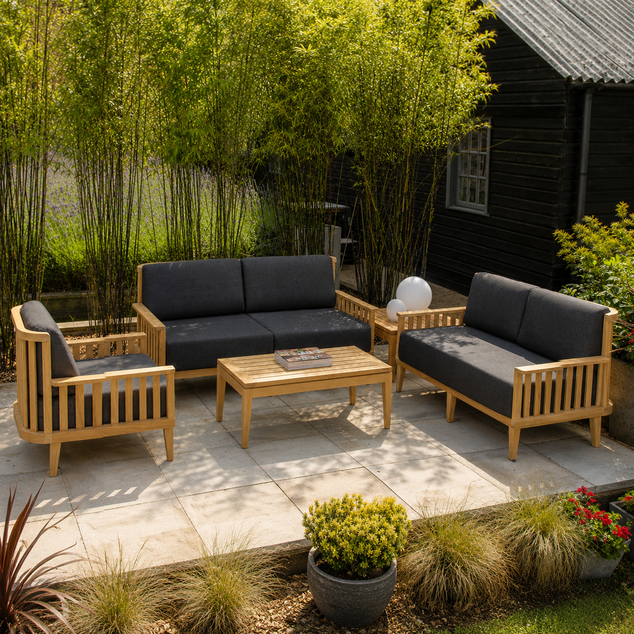 A 4-piece outdoor patio furniture set with light natural wood frames and dark charcoal gray cushions, arranged on a stone patio in a garden setting with bamboo and a black wooden cabin in the background.