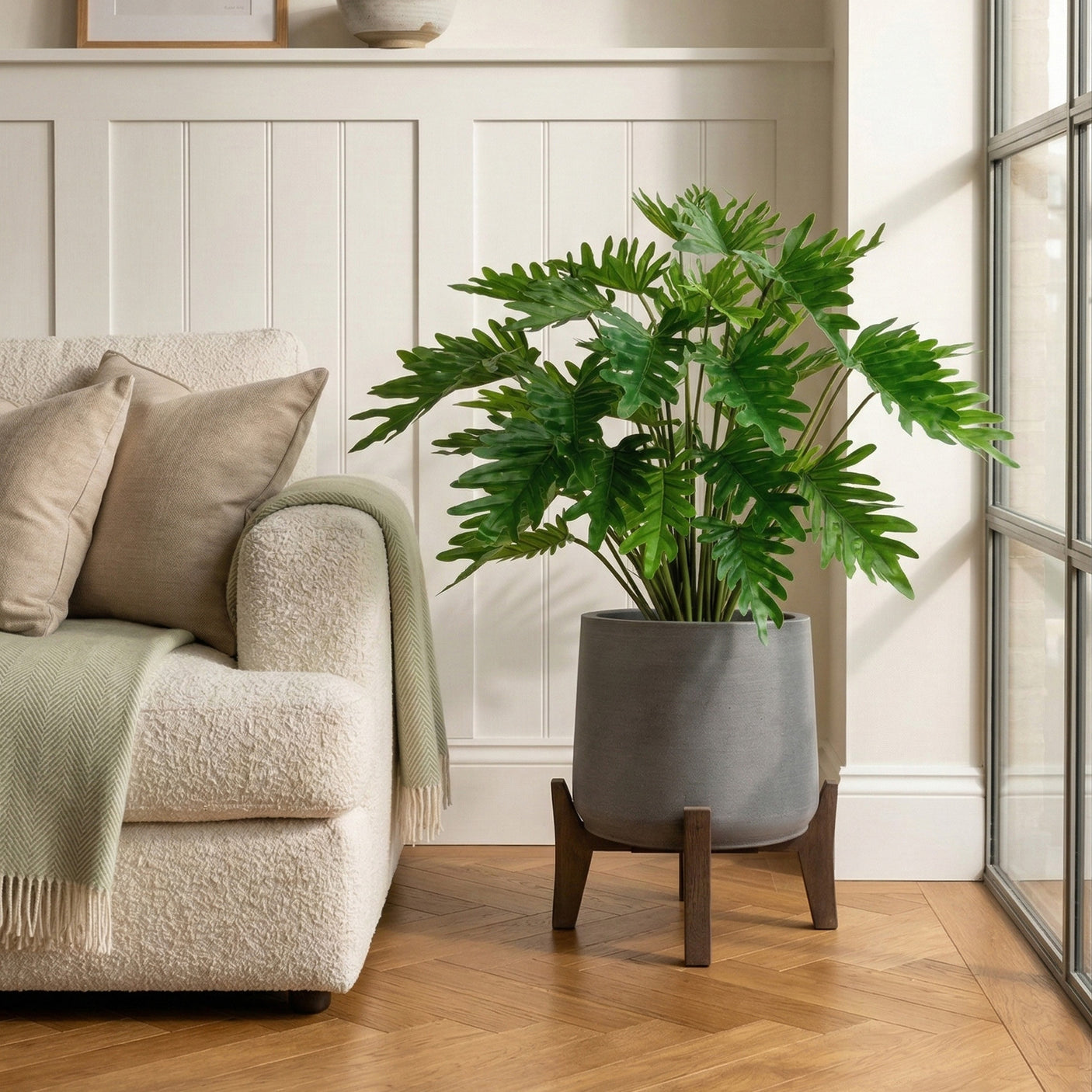 Living room with a beige sofa, green plant in a gray pot on a wooden stand, and white paneled walls.