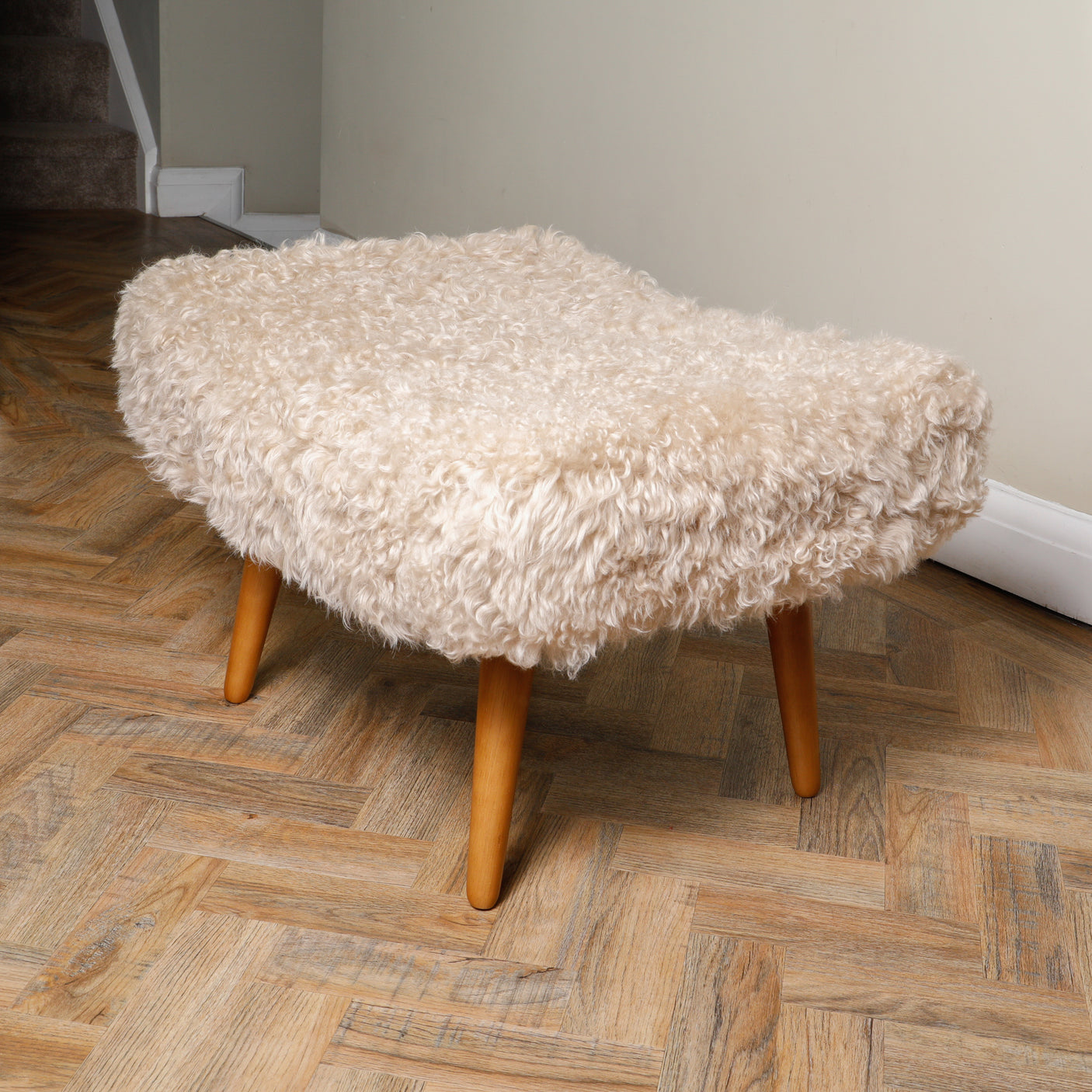 A beige shearling sheepskin footstool with dark oak stained legs on a wooden floor.