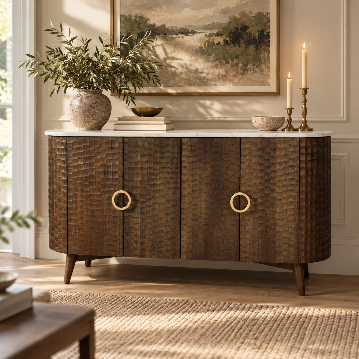 A mid-century modern sideboard cabinet with a dark brown textured wood finish, featuring two large circular brass handles and a white marble top. The cabinet is styled in a warm, sunlit room with a potted olive, candles, and a framed landscape painting.