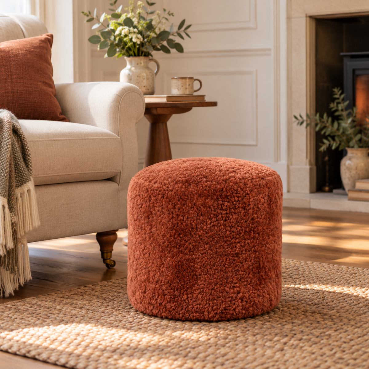 A round, textured terracotta sheepskin pouffe in a cosy living room setting next to a cream armchair and a fireplace.
