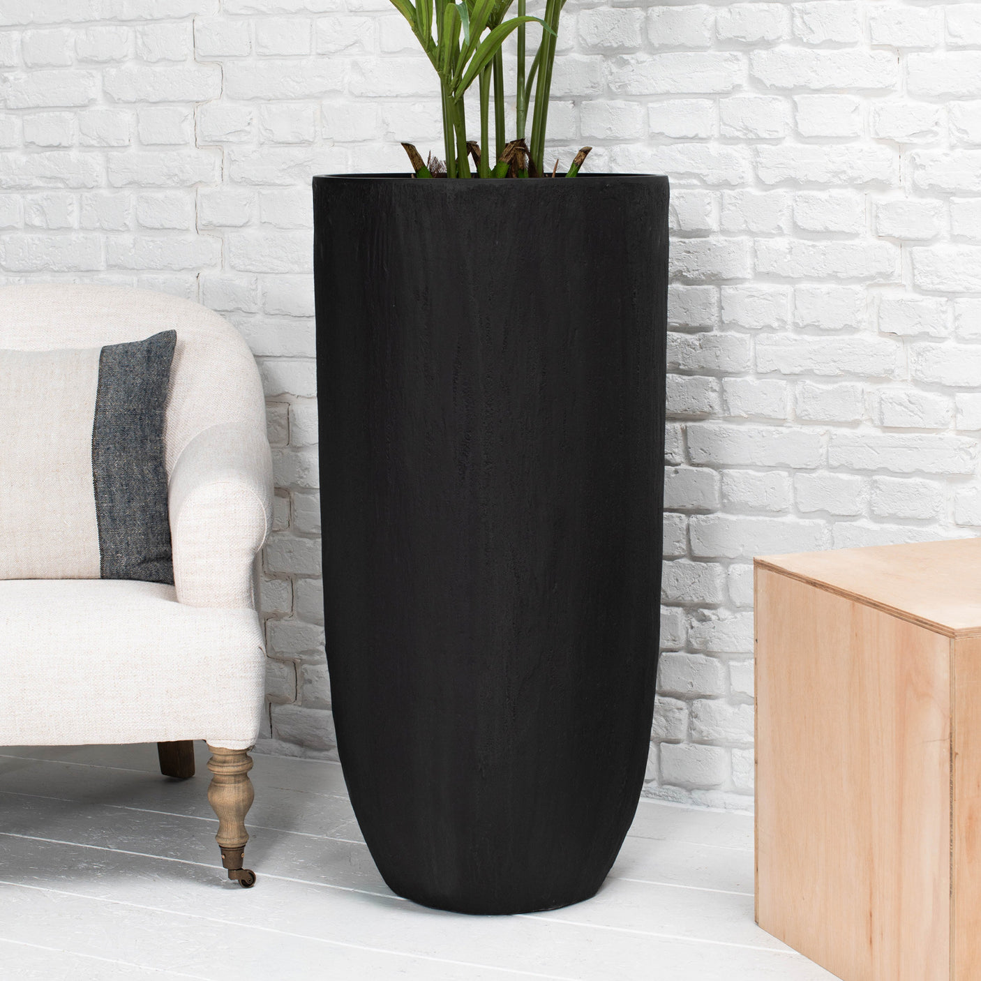 A tall, matte black cylindrical planter with a wood-grain texture, filled with green plant stems, set against a white brick wall in a styled interior scene.