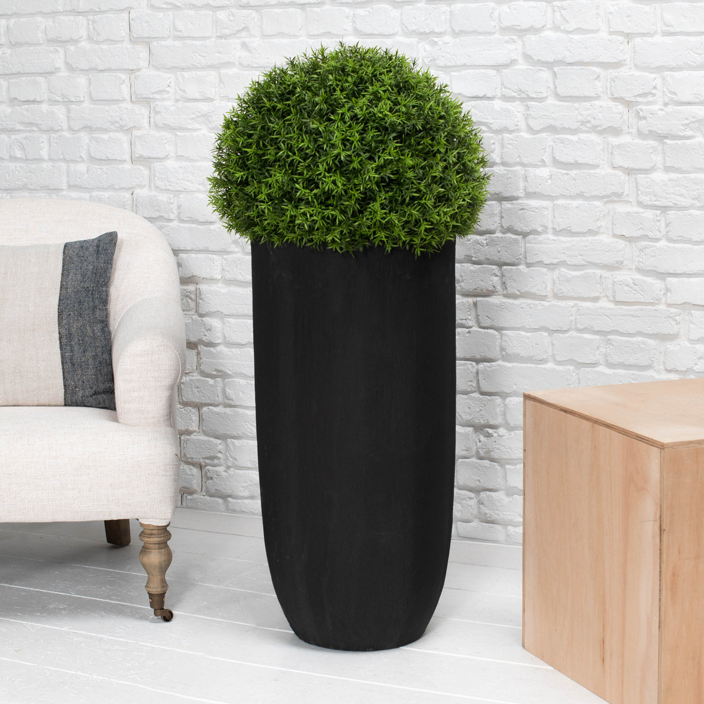 A tall black planter holding a rounded green topiary tree, styled in a living room setting with a white brick wall, a light-colored armchair, and a wooden side table.
