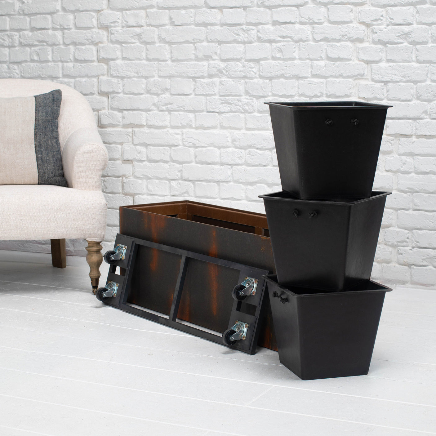 A black metal storage planter box with wheels, shown alongside three nested black square planters on a white brick floor next to a light-colored armchair.