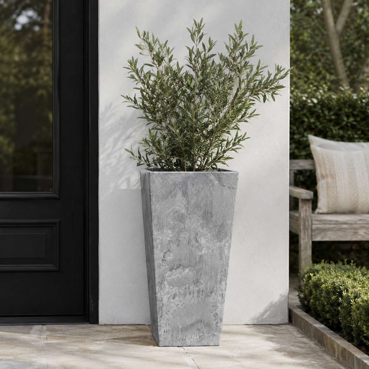 A tall grey outdoor planter against a white wall in an outdoor setting beside a black front door