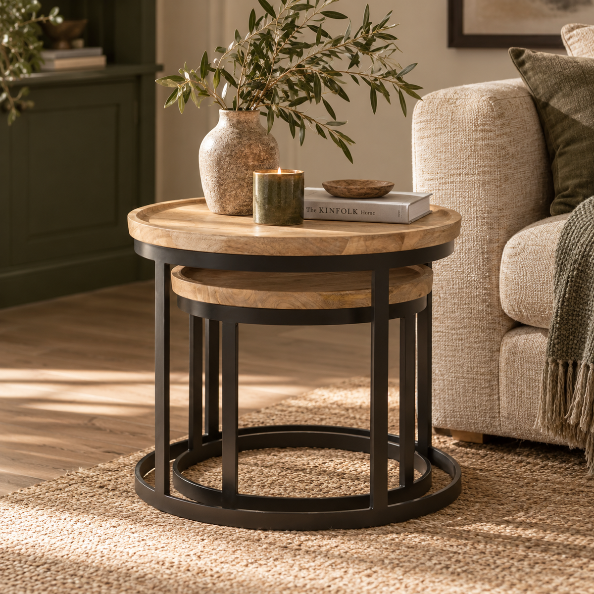 A round nesting side table with a light oak wood top and black metal frame, styled in a cosy living room next to a beige sofa with a vase and candle on top.