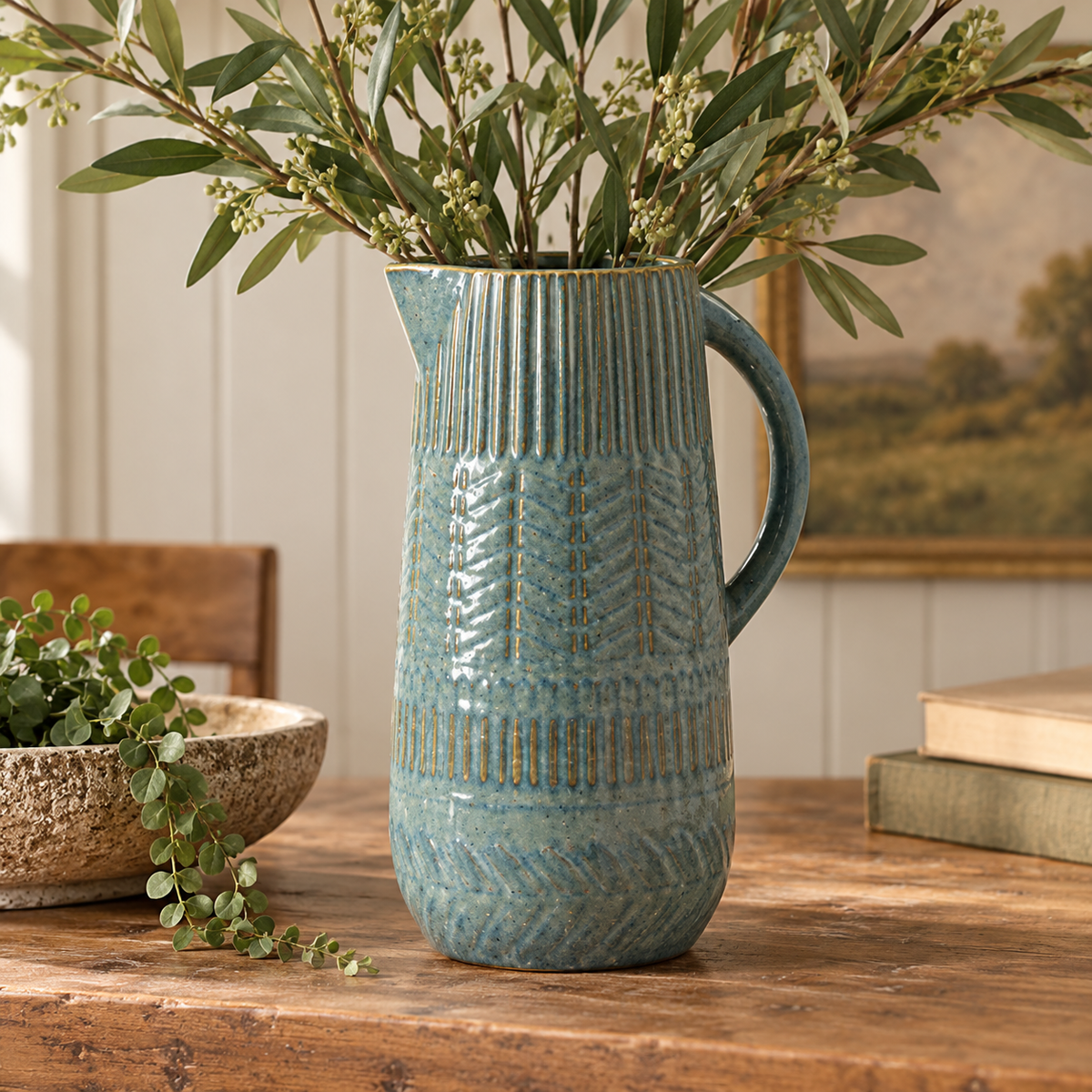 Decorative blue ceramic jug with textured detailing, styled with olive branches on a rustic wooden table in a warm country-style interior.