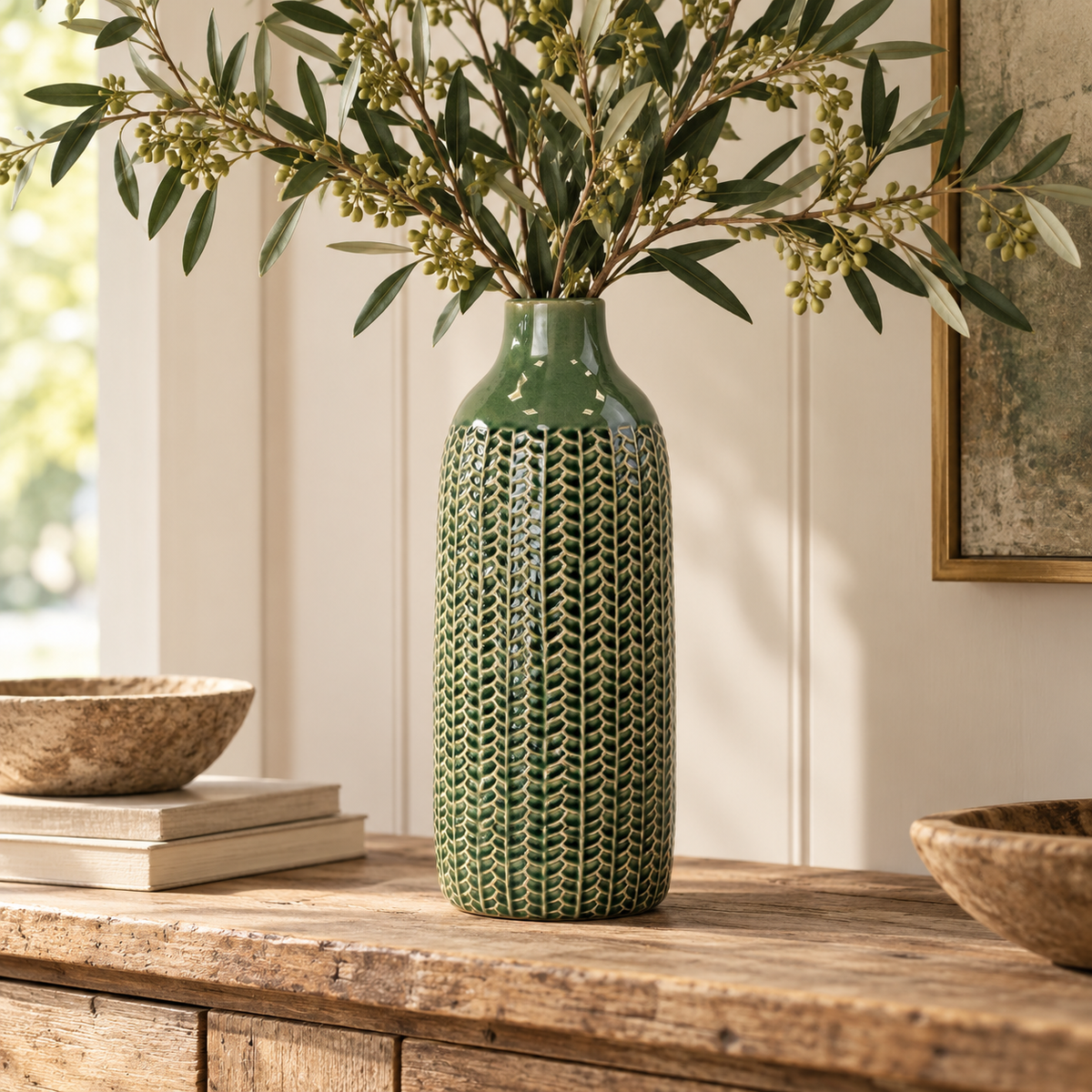 Tall deep green textured ceramic vase with olive branches displayed on a rustic wooden console in a warm, neutral country-style interior.