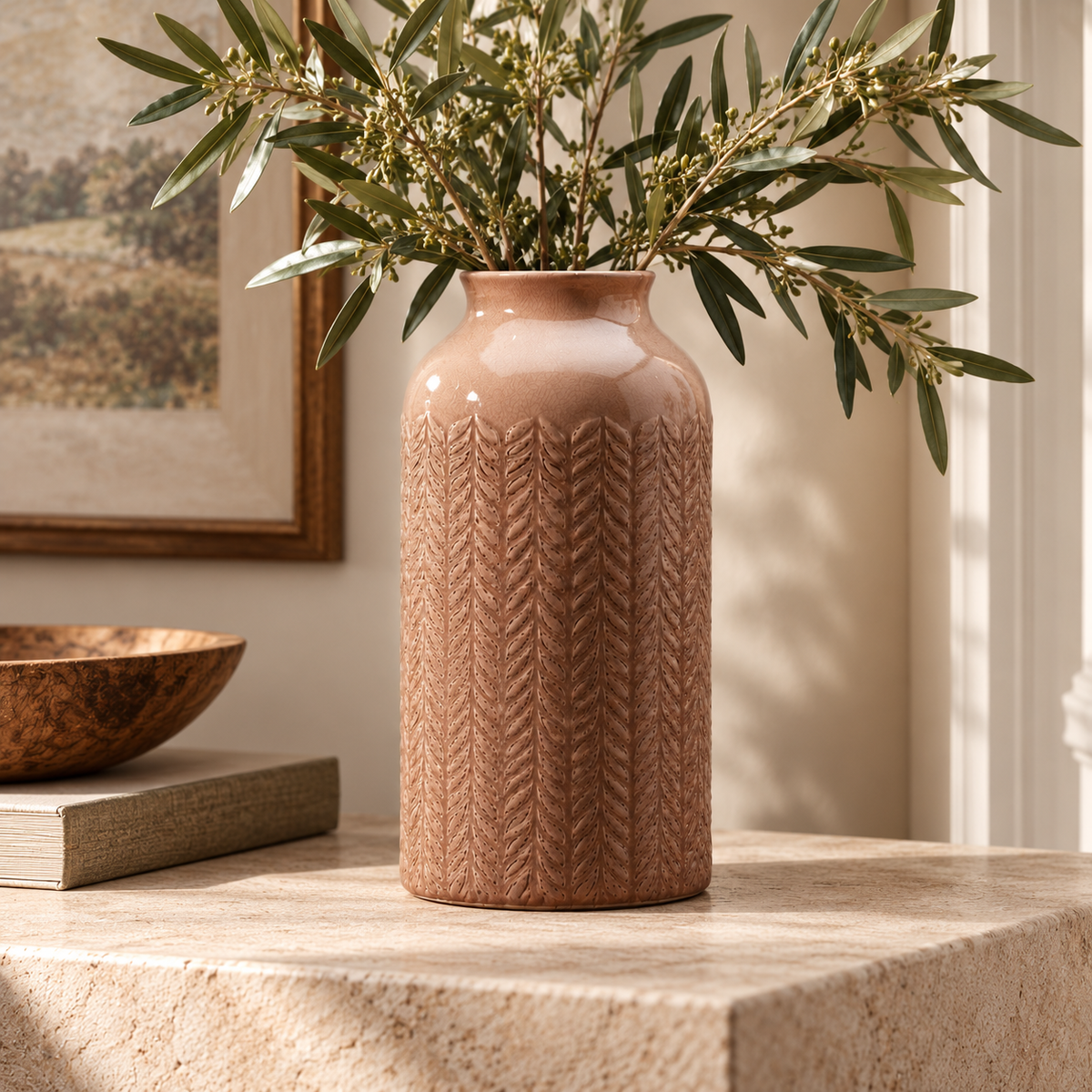 Dusky pink ceramic vase with carved leaf pattern filled with olive branches, styled on a natural stone console in a warm country-style interior.