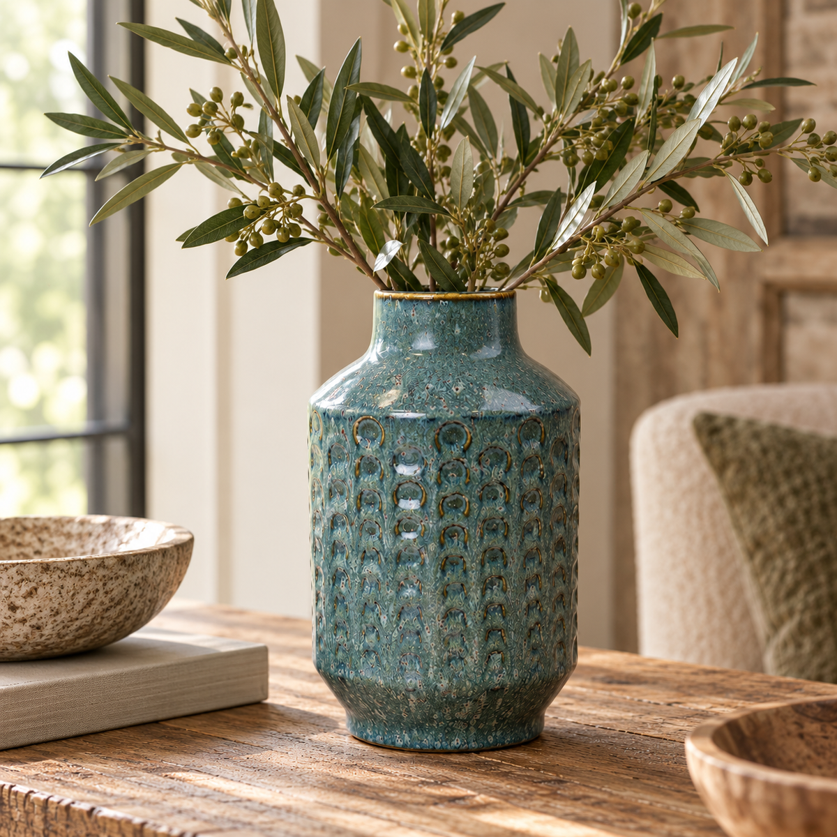 Teal ceramic vase with a textured embossed pattern and speckled glaze, styled with olive branches on a rustic wooden table in a warm, neutral-toned interior.