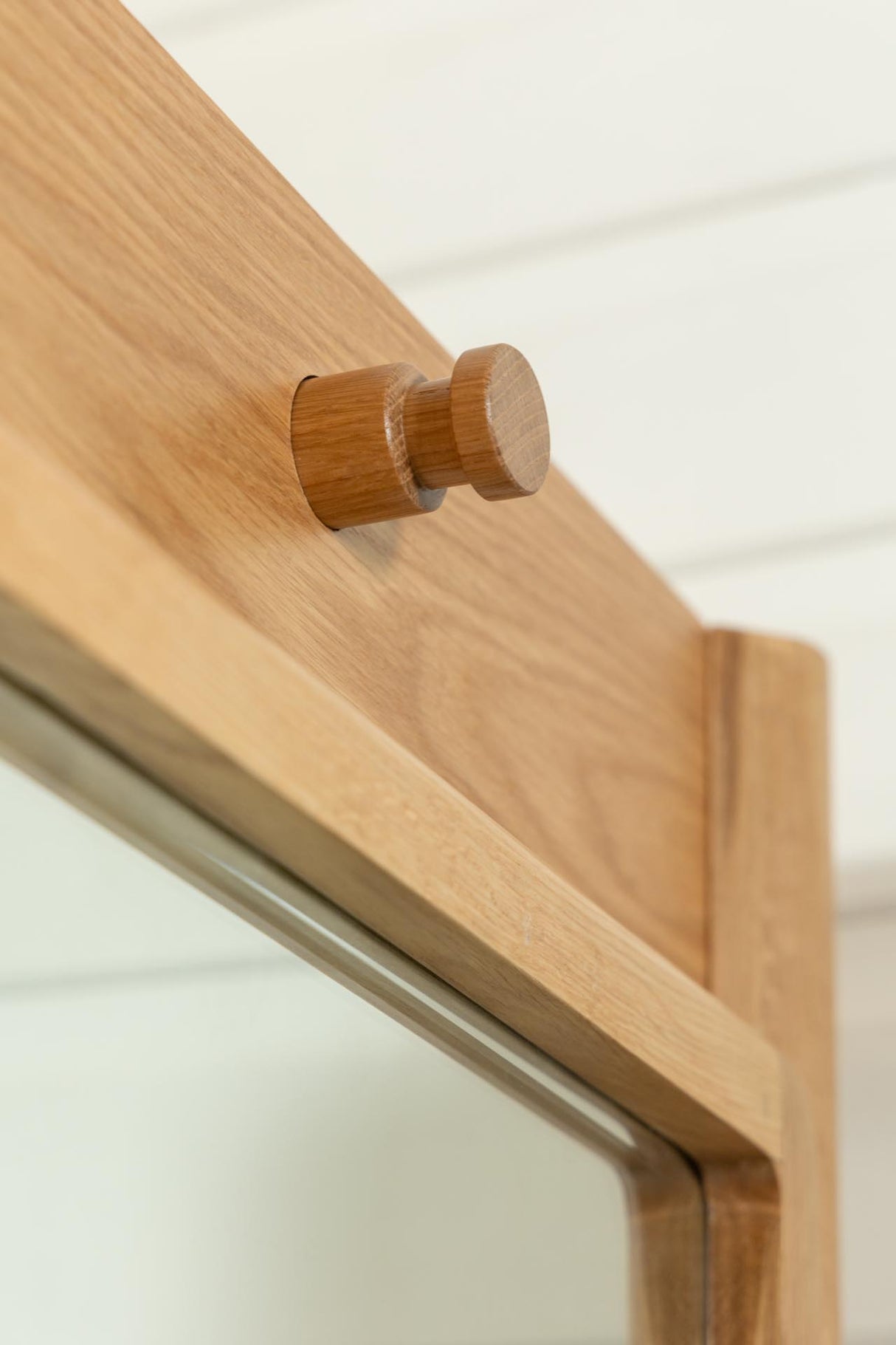 Close-up of a light natural wood cabinet knob mounted on a wooden cabinet door, with a glass panel and white paneled wall in the background.