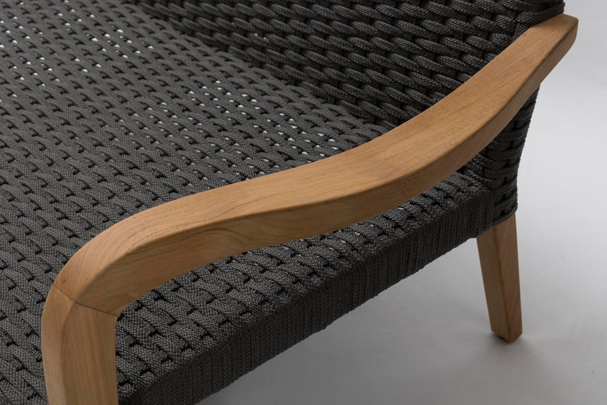 Close-up of a woven outdoor sofa showing dark charcoal grey rope upholstery, a curved natural wood armrest, and light wood legs on a light grey floor.