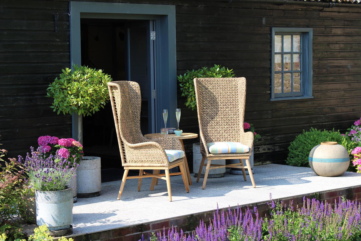 Two tall woven wicker patio chairs with light wood legs and striped cushions sit on a stone patio in front of a dark wood house, with potted plants and flowers around them.