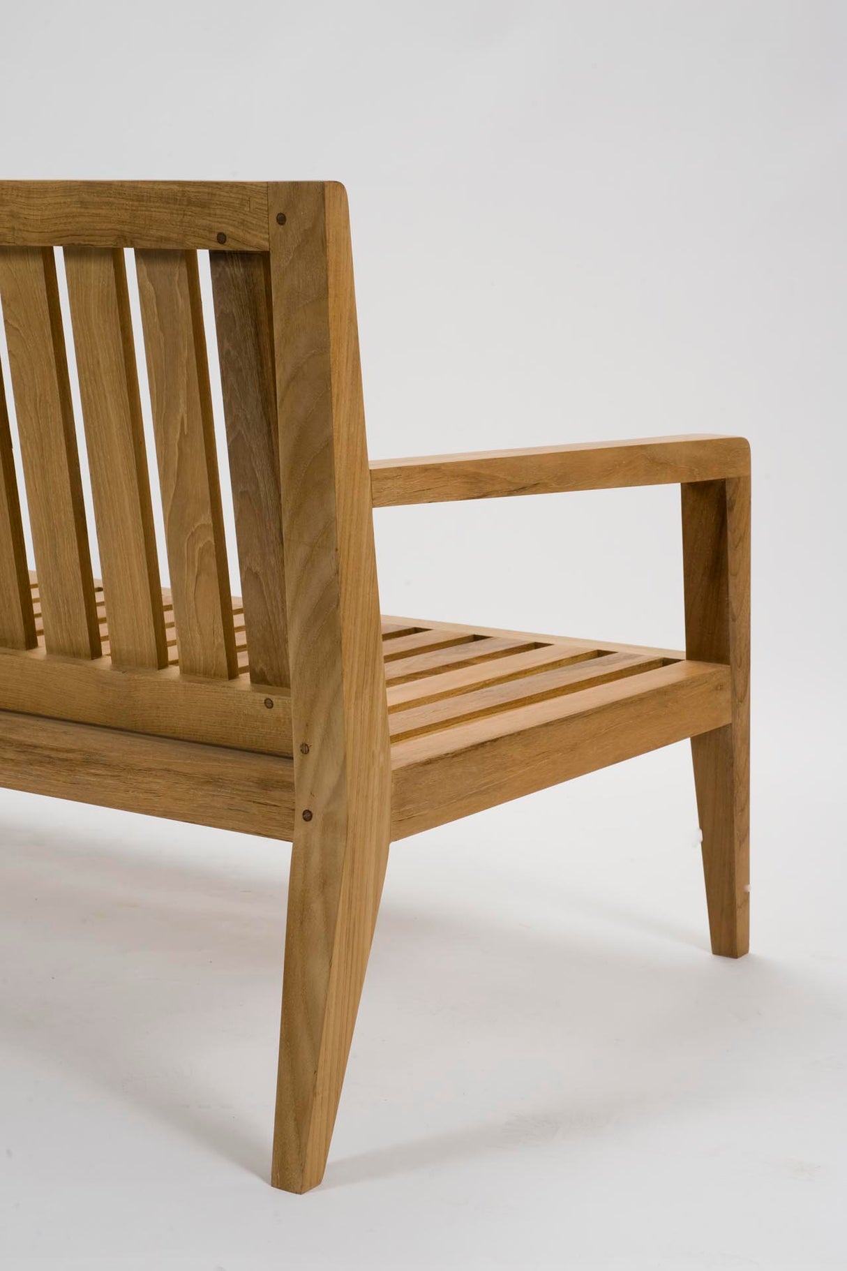 Close-up of a wooden outdoor bench showing the backrest, armrest, slatted seat, and angled legs in a natural light brown wood finish against a white studio background.