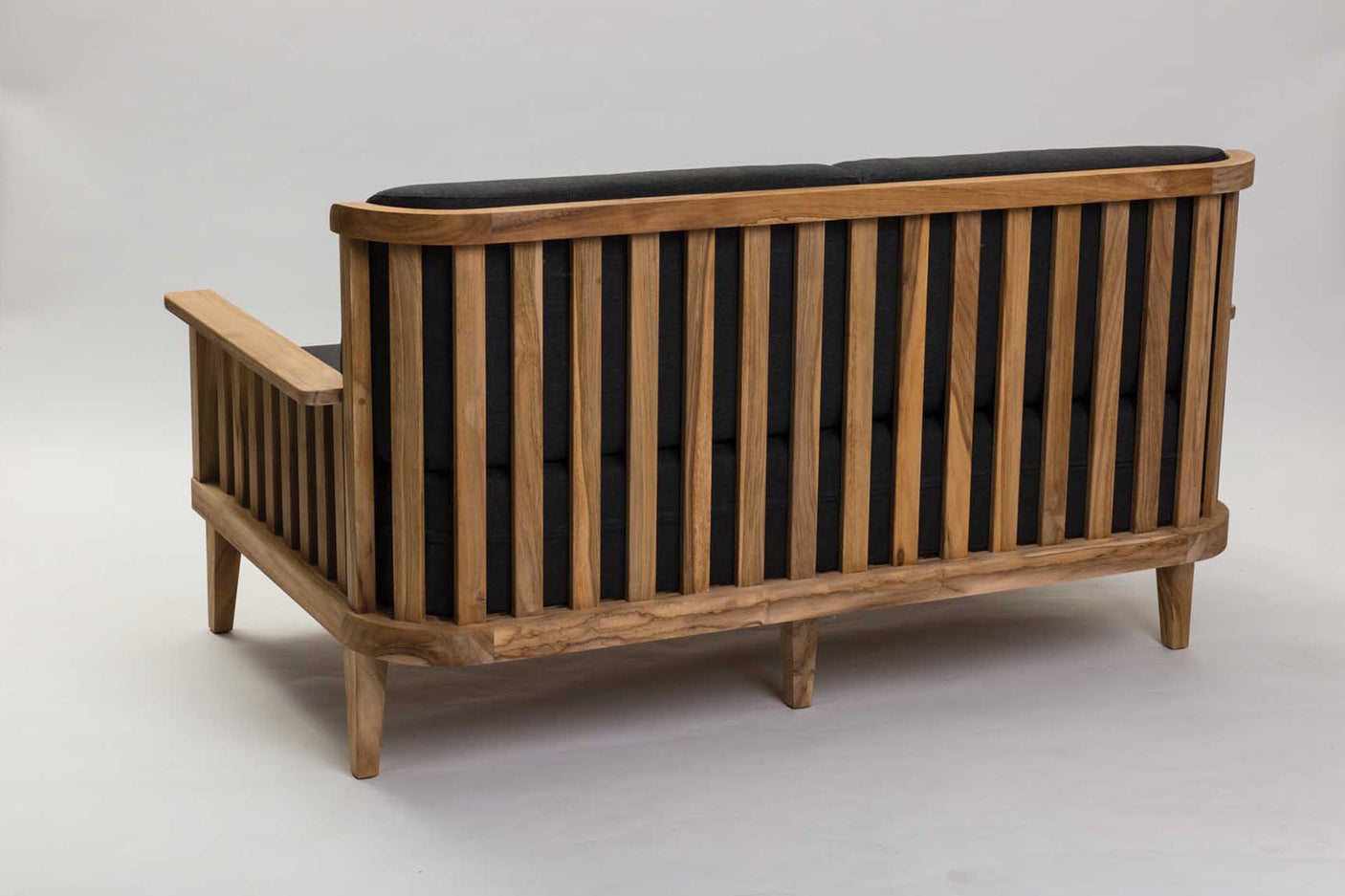 A mid-century modern wooden outdoor sofa with a slatted backrest and armrests, featuring a natural oak wood frame and a black cushioned seat and back, photographed on a seamless light gray studio background.
