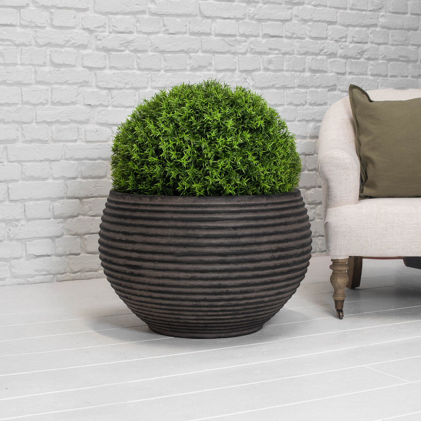 A spherical green topiary tree in a dark brown ribbed ceramic pot, styled next to a light beige armchair against a white brick wall background.