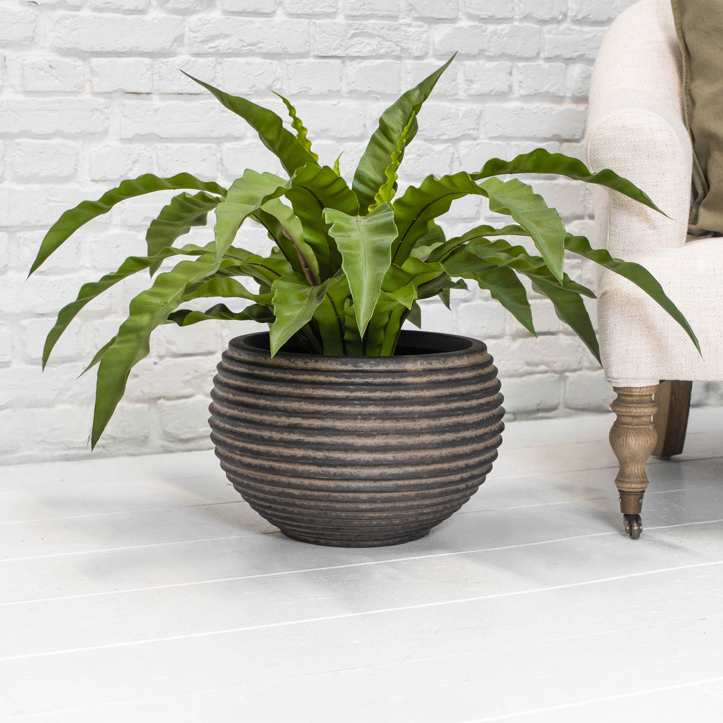 A round ribbed brown planter with a green plant on a light grey floor next to a chair in front of a white brick wall