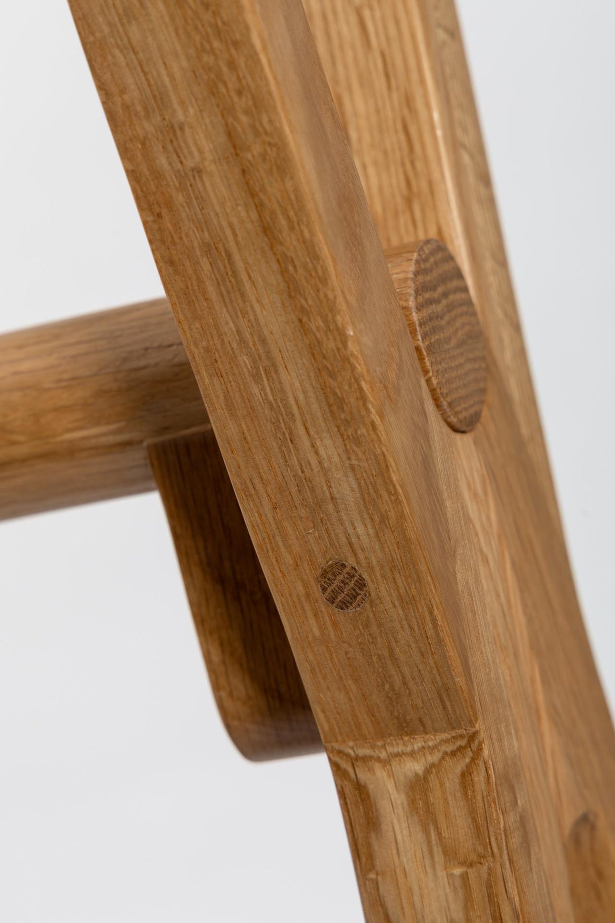 Close-up detail of a wooden chair or stool leg joint, showing natural wood grain, a circular peg, and a dowel on a light gray background.