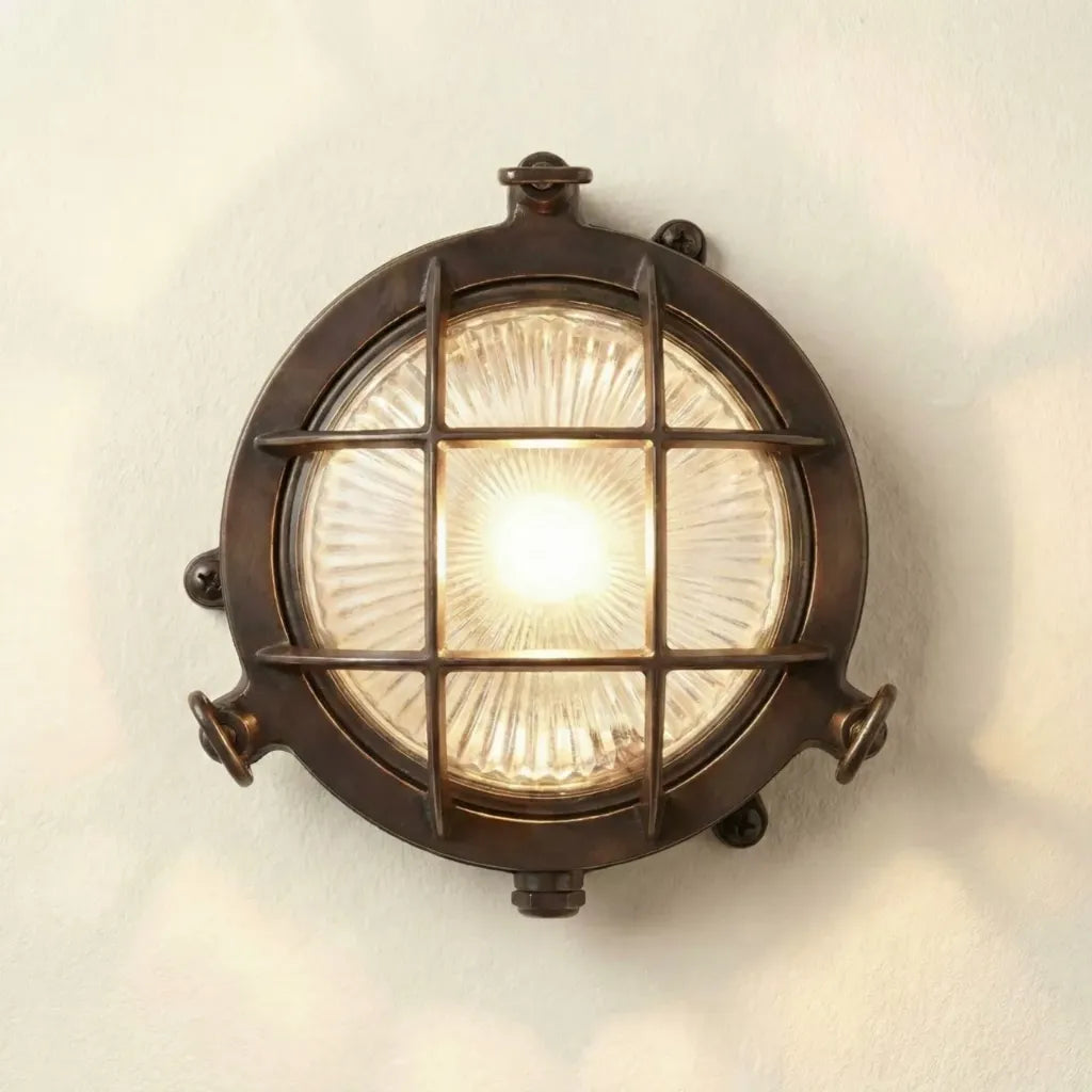 A round nautical-style wall light fixture with a dark bronze metal frame, protective wire grid, and a glowing clear glass shade, mounted on a plain off-white wall.