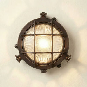 A round nautical-style wall light fixture with a dark bronze metal frame, protective wire grid, and a glowing clear glass shade, mounted on a plain off-white wall.