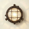 A round nautical-style wall light fixture with a dark bronze metal frame, protective wire grid, and a glowing clear glass shade, mounted on a plain off-white wall.