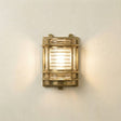 A rectangular industrial wall sconce with a brass-finished metal frame and ribbed clear glass panels, mounted on a textured off-white wall and casting a warm light.