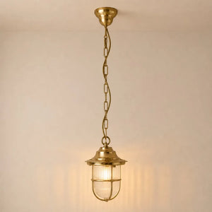 A hanging pendant light fixture with a warm brass finish, featuring a ribbed glass shade and a safety cage, suspended by a chain against a plain wall.