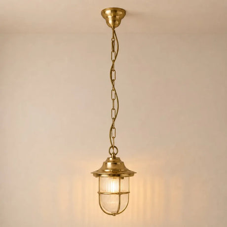 A hanging pendant light fixture with a warm brass finish, featuring a ribbed glass shade and a safety cage, suspended by a chain against a plain wall.