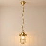 A hanging pendant light fixture with a warm brass finish, featuring a ribbed glass shade and a safety cage, suspended by a chain against a plain wall.