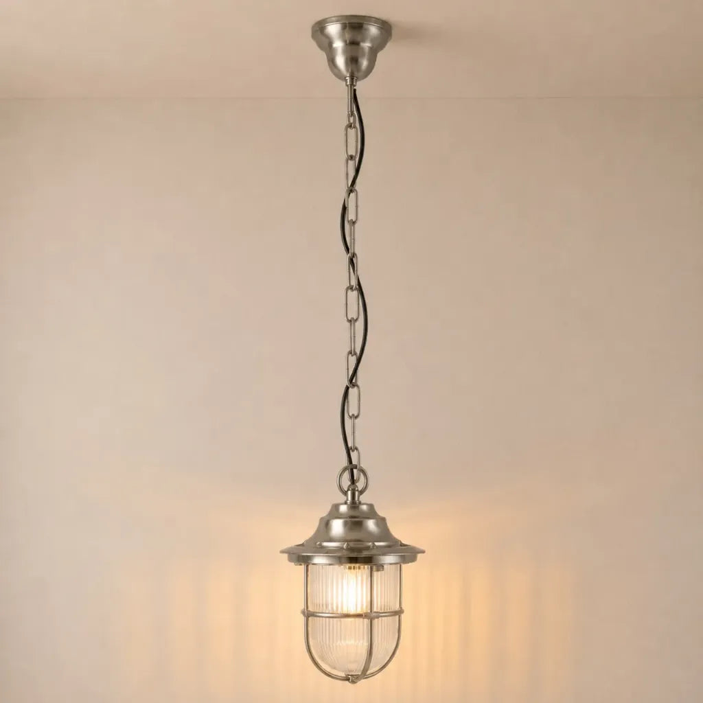 A hanging pendant light with a brushed silver metal finish, featuring a ribbed glass shade and a wire safety cage, suspended by a chain against a warm beige wall.
