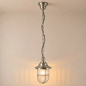 A hanging pendant light with a brushed silver metal finish, featuring a ribbed glass shade and a wire safety cage, suspended by a chain against a warm beige wall.