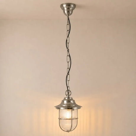 A hanging pendant light with a brushed silver metal finish, featuring a ribbed glass shade and a wire safety cage, suspended by a chain against a warm beige wall.