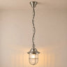 A hanging pendant light with a brushed silver metal finish, featuring a ribbed glass shade and a wire safety cage, suspended by a chain against a warm beige wall.